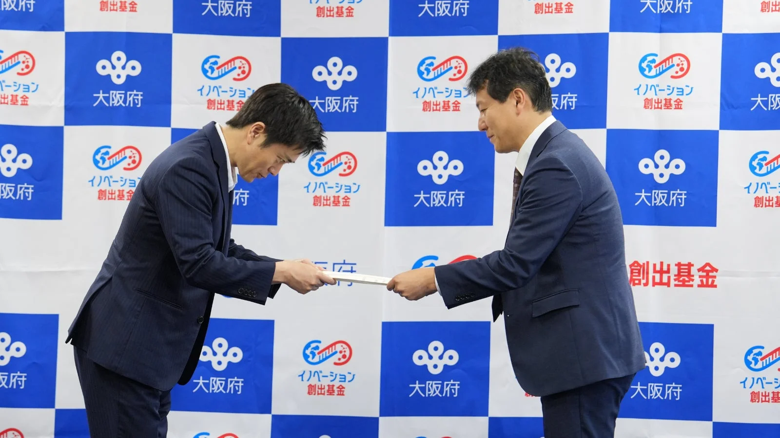 大阪府とイノベーション創出基金のロゴが描かれた背景の前で、二人のビジネスマンが丁寧にお辞儀をしながら書類を交換している場面