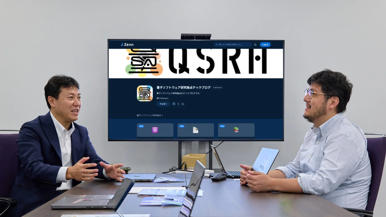 会議室で二人のビジネスマンが向かい合って話しており、背景の大型スクリーンには「量子ソフトウェア研究拠点テックブログ」のZennページが表示されています。