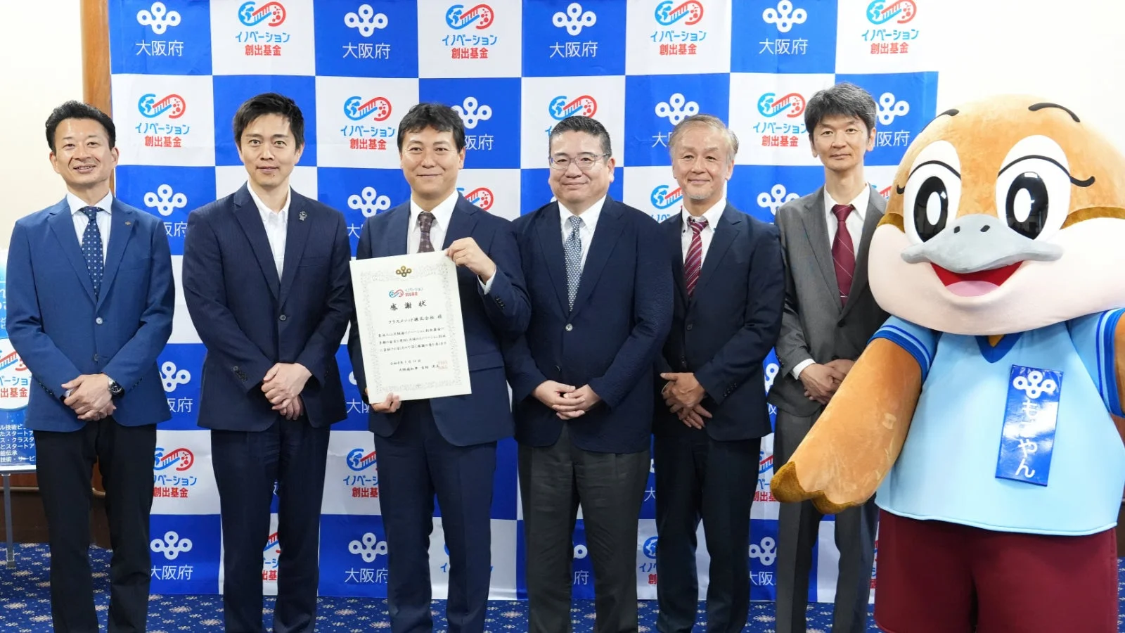 大阪府とイノベーション創出基金のロゴが入った背景の前で、6人の男性と大阪府のマスコット「もずやん」が集合写真を撮っています。中央の男性は感謝状を手にしています。
