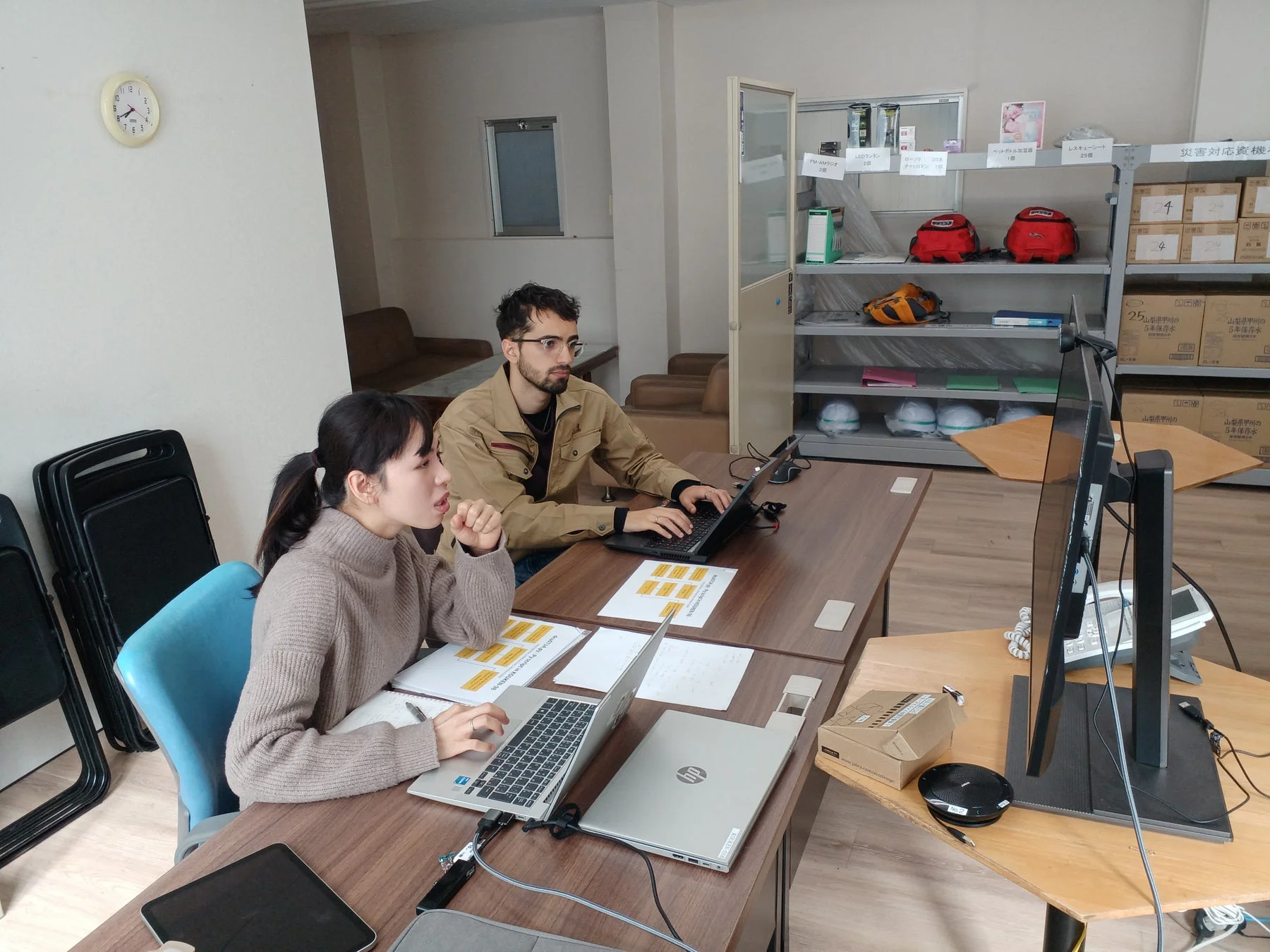 オフィスでノートパソコンに向かう男女。災害対策用品が並ぶ棚を背景に