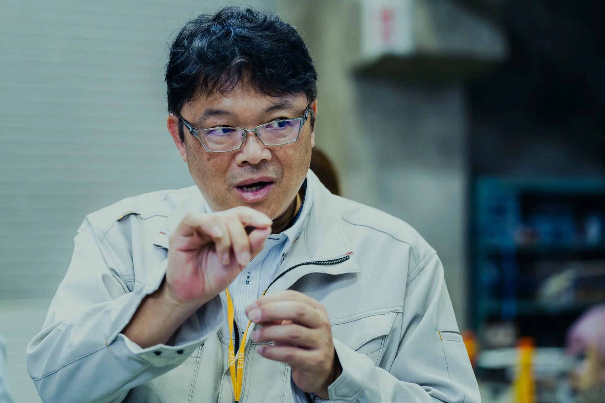 眼鏡をかけた中年の日本人男性が、作業着を着用し、真剣な表情で何かを話しながら手でジェスチャーをしている