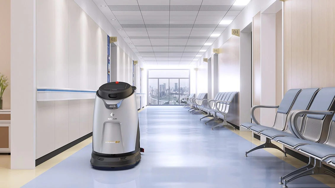 モダンな病院の廊下で、自律型清掃ロボットが稼働している様子です。廊下には待合用の椅子が並び、窓からは都市のビル群が見えます。清潔感のある空間で、医療施設の効率化を示唆しています。