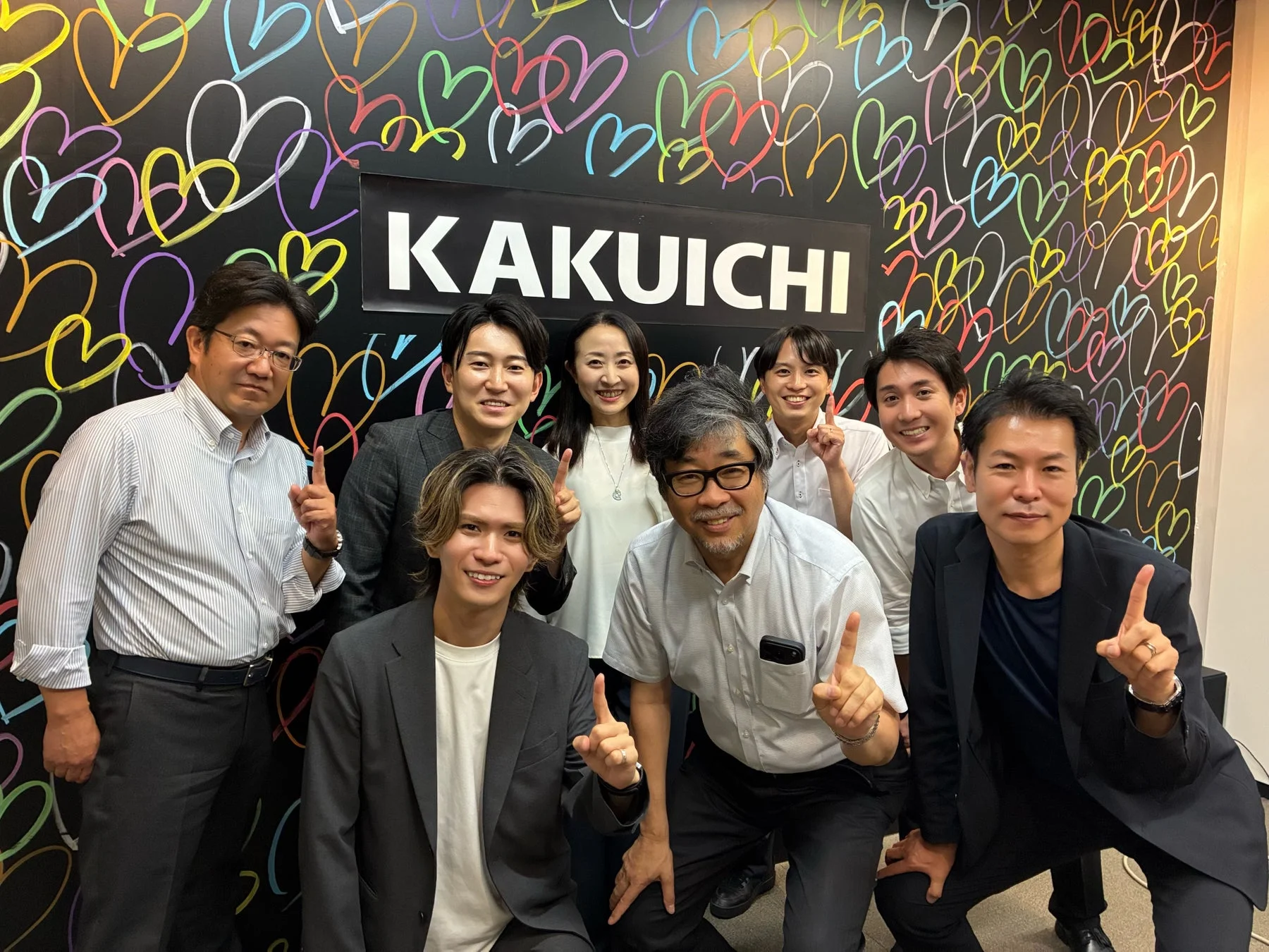 「KAKUICHI」のロゴとカラフルなハートが描かれた壁の前で、数名の男女が笑顔でポーズをとっている集合写真です。親指を立てたり指を差したりして、和やかな雰囲気が伝わります。