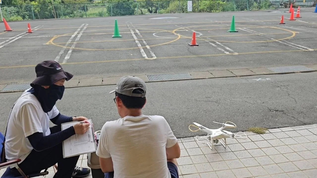 屋外の舗ローンエリアでドローンの訓練か試験が行われている様子です。一人が記録を取り、もう一人がドローンとコースを観察しており、地面には白いドローンが置かれ、背景にはコーンとラインが設置されています。