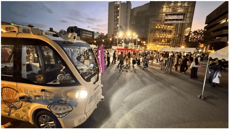 夕暮れ時の屋外イベントで、多くの人々が集まる中に自動運転バス