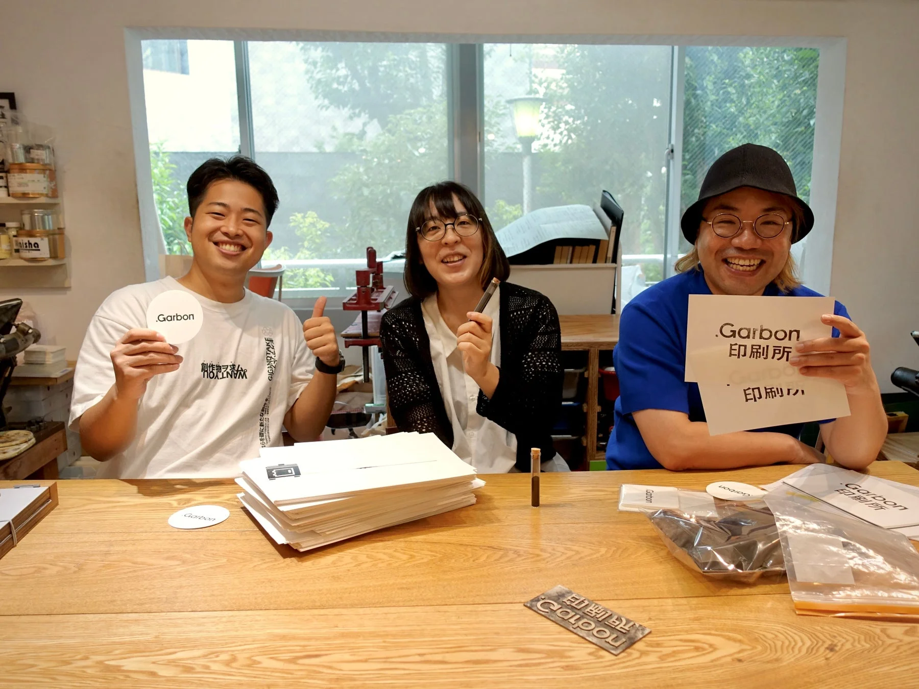 笑顔の男女3人が木製のテーブルに座り、「.Garbon」や「印刷所」と書かれた紙や製品を見せている。