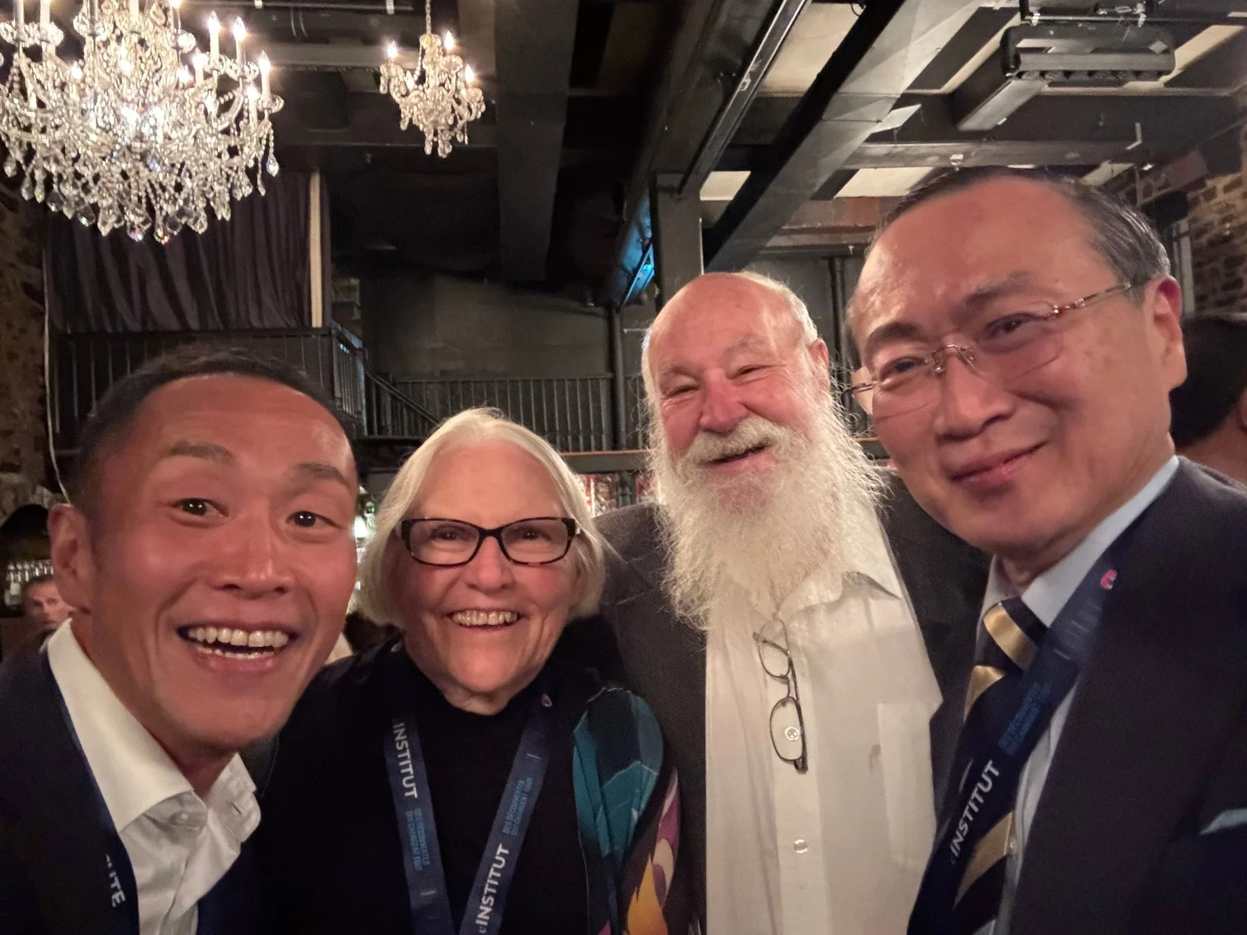 4人の男女が笑顔でセルフィーを撮っている写真。背景にはシャンデリアが見え、何らかのイベントや会議の場での交流の様子を捉えている。