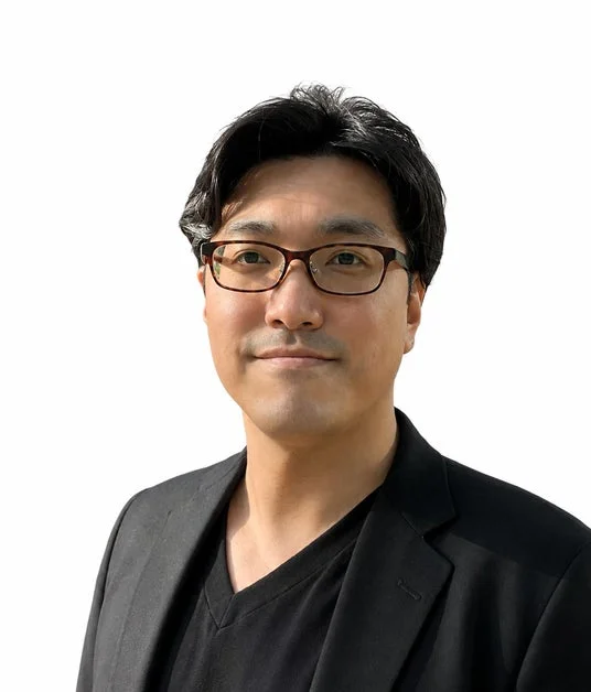 眼鏡をかけたアジア系の男性が、黒いジャケットとVネックシャツを着用し、カメラを見ている上半身のポートレート