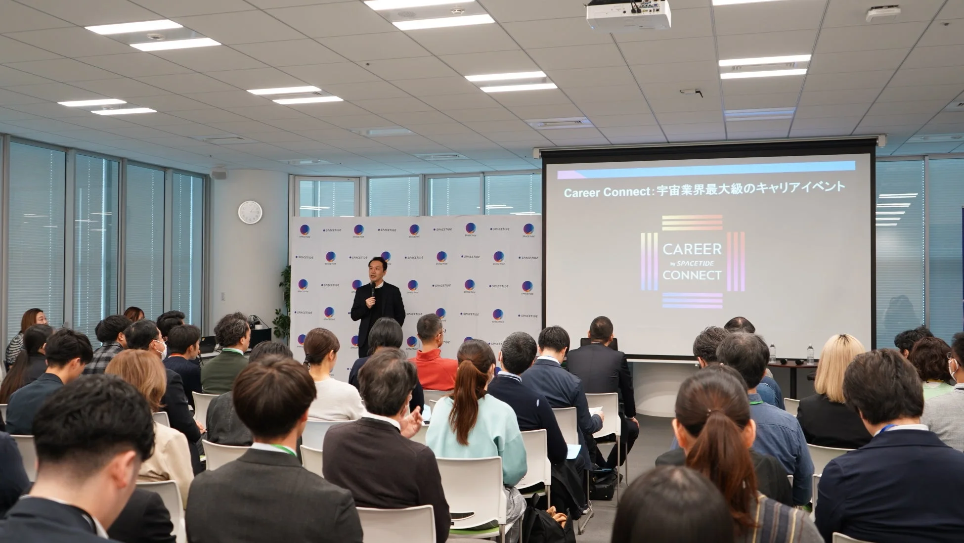 企業イベント会場で男性が講演しており、多くの聴衆が耳を傾けている