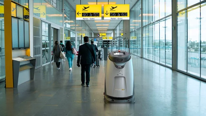 空港のターミナル内を数人の人々が歩いており、自律走行式の清掃ロボットが稼働している様子です。頭上にはゲートへの方向を示す標識があり、大きな窓からは外の景色が見えます。