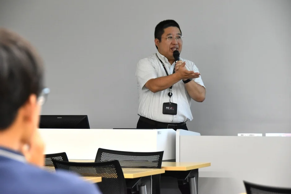 会議室で男性がマイクを持ってプレゼンテーションを行っており、聴講者が熱心に耳を傾けている様子。