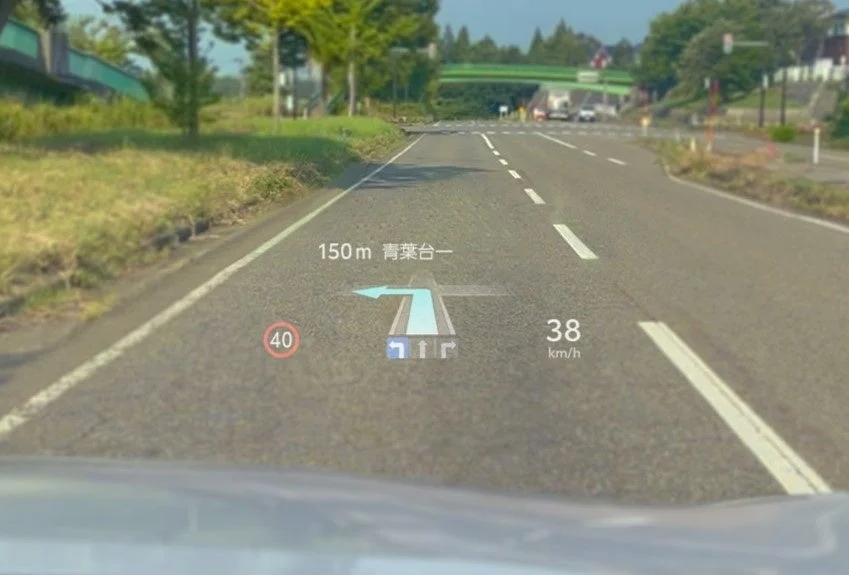 車のフロントガラスに表示されたヘッドアップディスプレイ（HUD）の映像。150m先で青葉台方面へ左折するナビゲーション指示、現在の速度、制限速度が表示