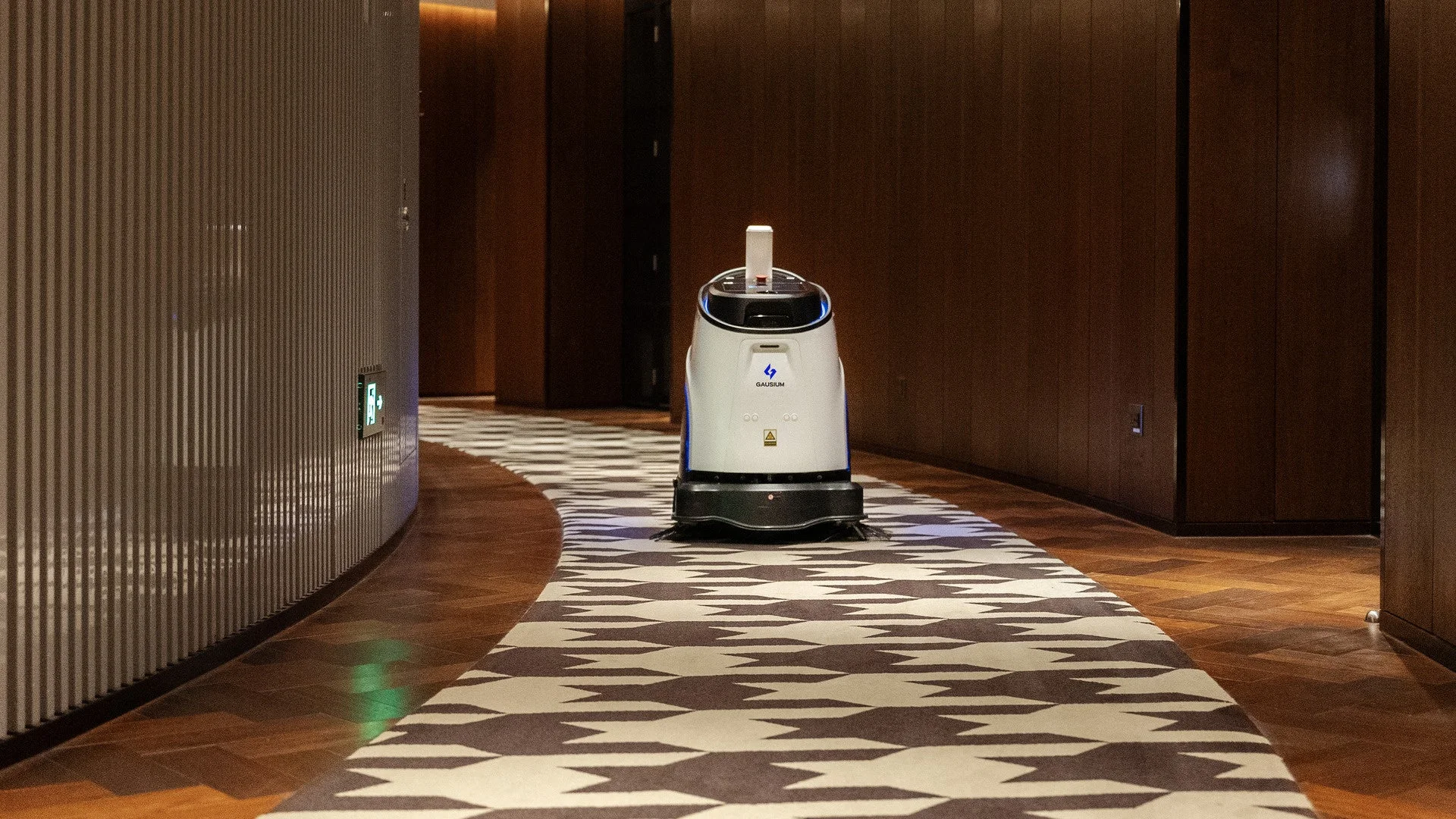 モダンな内装のホテルまたは商業施設の廊下で、自動清掃ロボットが特徴的な柄のカーペット上を走行している様子です。
