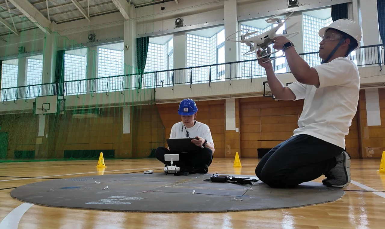体育館でドローンの操作訓練を行っている様子。一人がドローンを持ち、もう一人がタブレットで操作設定をしている。