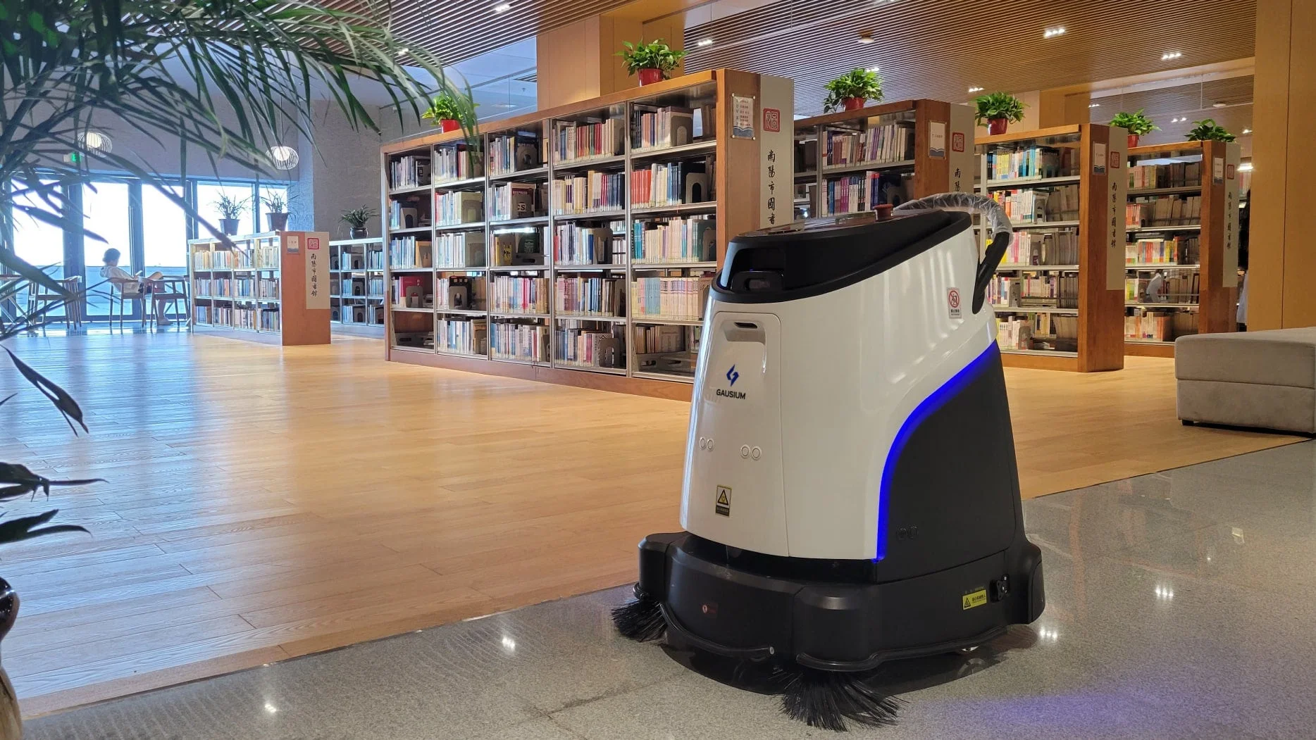モダンな図書館の内部で、自動清掃ロボットが稼働している様子です。奥には書架が並び、窓際には読書をする人が見えます。清潔感のある広々とした空間です。