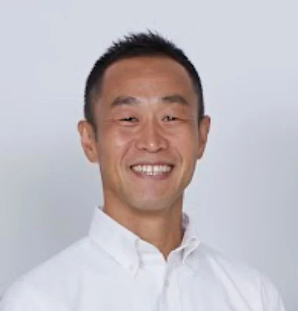 白い背景の前で白いシャツを着た短髪の日本人男性が、明るく笑顔を見せているポートレート写真。