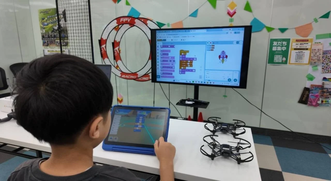 子供がタブレットとモニターを使ってブロックプログラミングに取り組んでいます。テーブルには2台のドローンがあり、プログラミング学習を通じてドローン操作などを学んでいる様子が伺えます。