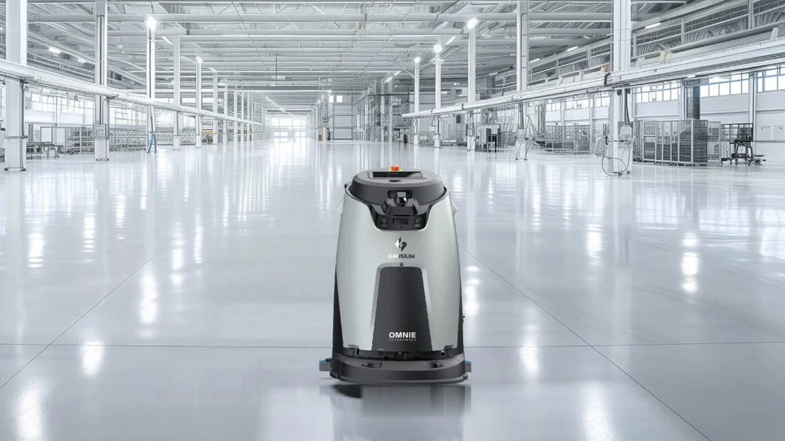 広々とした明るい工場または倉庫の床の中央に、銀色と黒の自律型清掃ロボットが置かれています。床は非常にきれいで光沢があり、ロボットがその清潔さを維持している様子が伺えます。