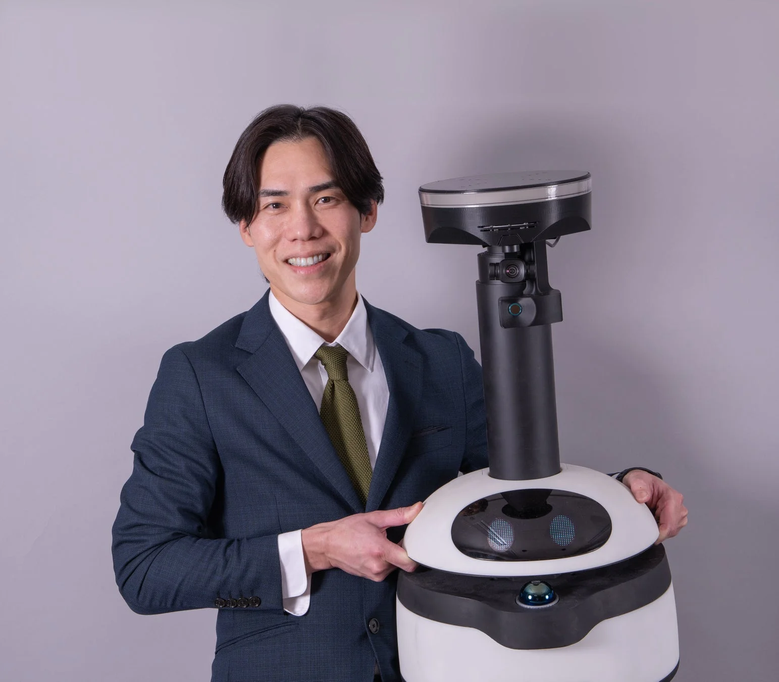 ビジネススーツを着た男性が、白いサービスロボットを笑顔で抱えている画像です。男性はカメラ目線で、ロボットとの親和性やテクノロジーの共存を表現しています。