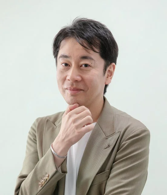 ベージュのスーツジャケットと白Tシャツを着た男性が、顎に手を当ててカメラに優しく微笑んでいます