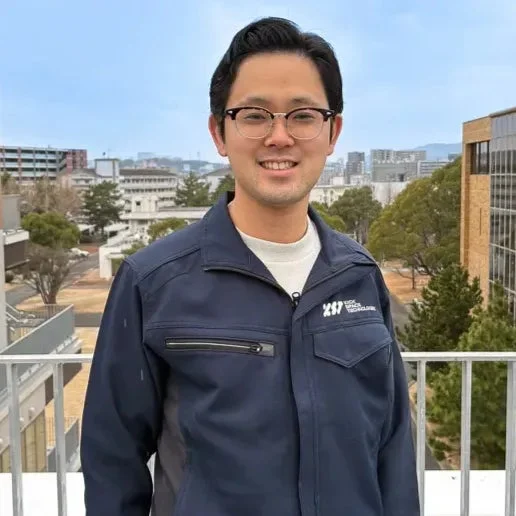 紺色の作業服を着て眼鏡をかけたアジア人男性が、屋外の建物が立ち並ぶ場所で笑顔を見せています。背景には青空が広がっています。