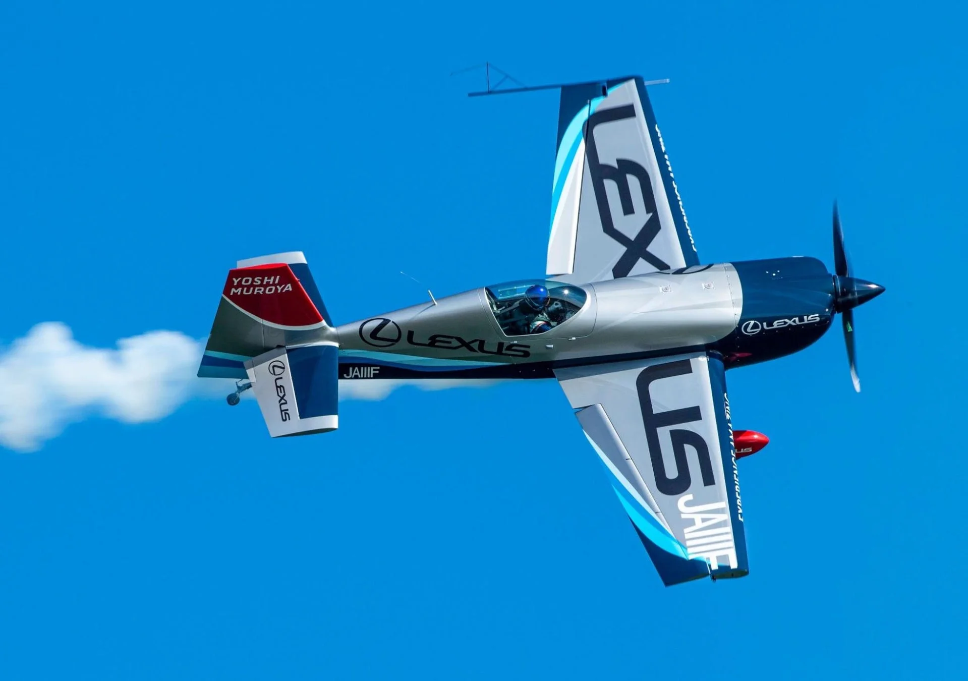 青い空を背景に、LEXUSのロゴと「YOSHI MUROYA」の名前が描かれた銀と青のアクロバット飛行機が、白いスモークを出しながら飛行している様子です。パイロットが操縦席にいるのが確認できます。