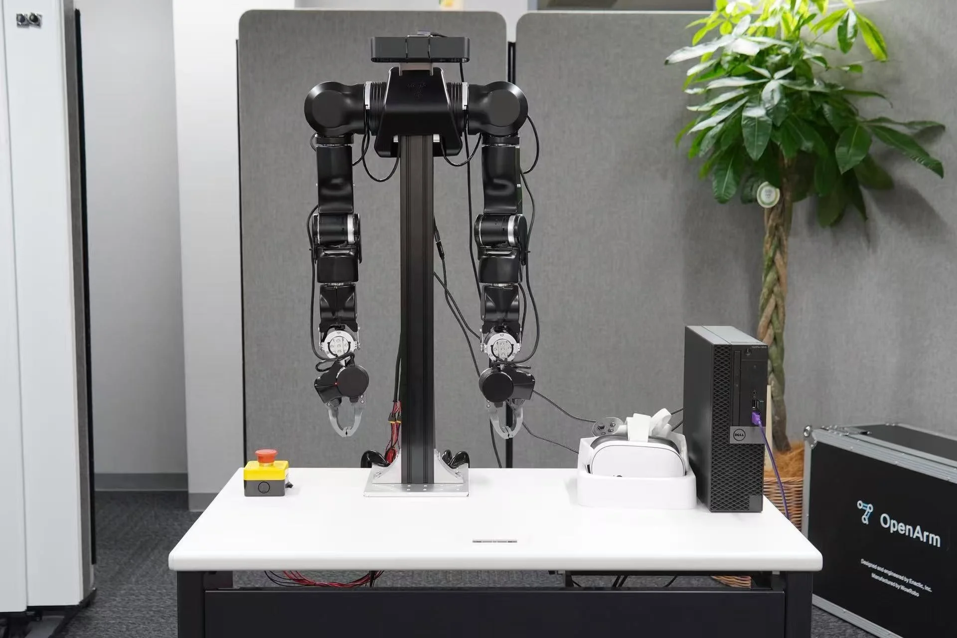 オフィス環境の白い机の上に設置された双腕ロボットシステムが写っています。PC、VRヘッドセット、緊急停止ボタンも配置されており、研究開発またはデモンストレーションの様子を示唆しています。