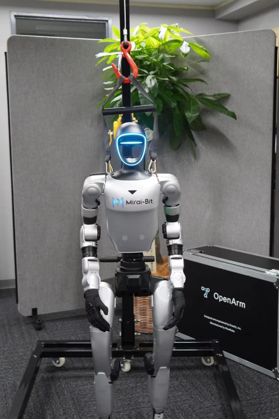 人型ロボット「Mirai-Bit」が吊り下げられた状態で展示されている様子。隣には「OpenArm」と書かれた黒いケースがあり、未来技術を感じさせる一枚です。